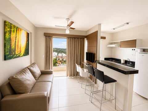 Modern studio apartment with beige sofa, kitchenette, bar seating, flat-screen TV, and balcony overlooking hillside town